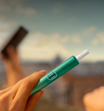 A woman holds an IQOS device while looking at a laptop.